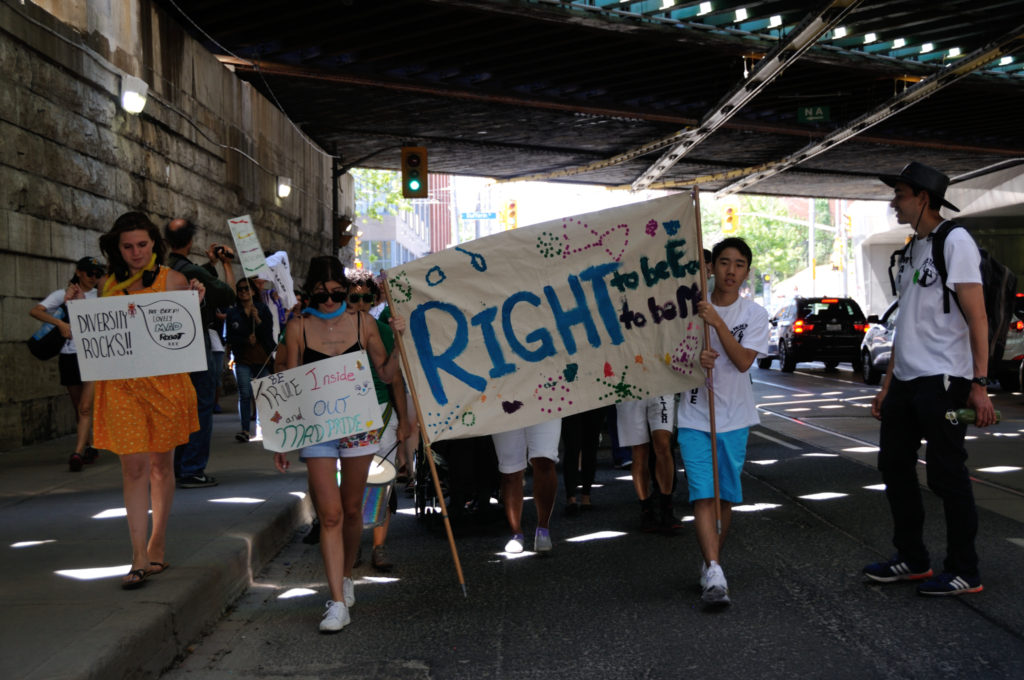Image: Marchers carrying signs like "Right to be free and Right to be me", walking under a bridge. Text: Right to be me Right to be free.,